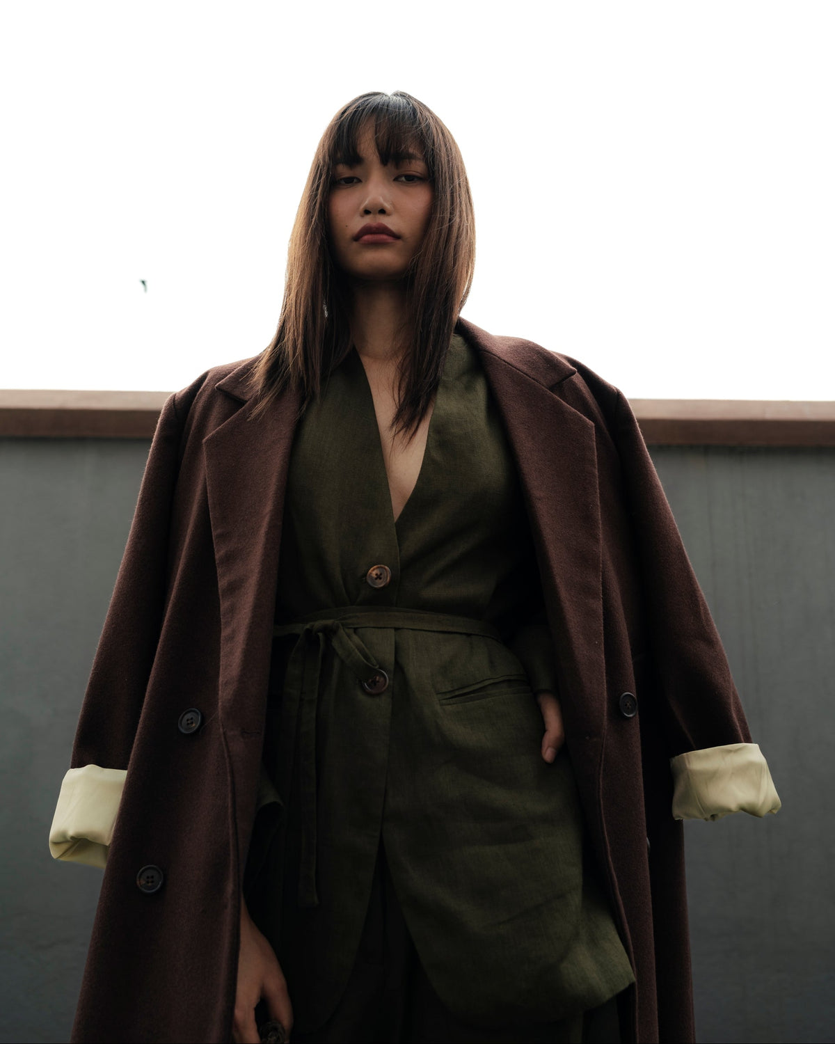 A woman wearing a dark brown double-breasted coat with cream turned-up cuffs over an olive green belted dress, standing against a high white sky background.