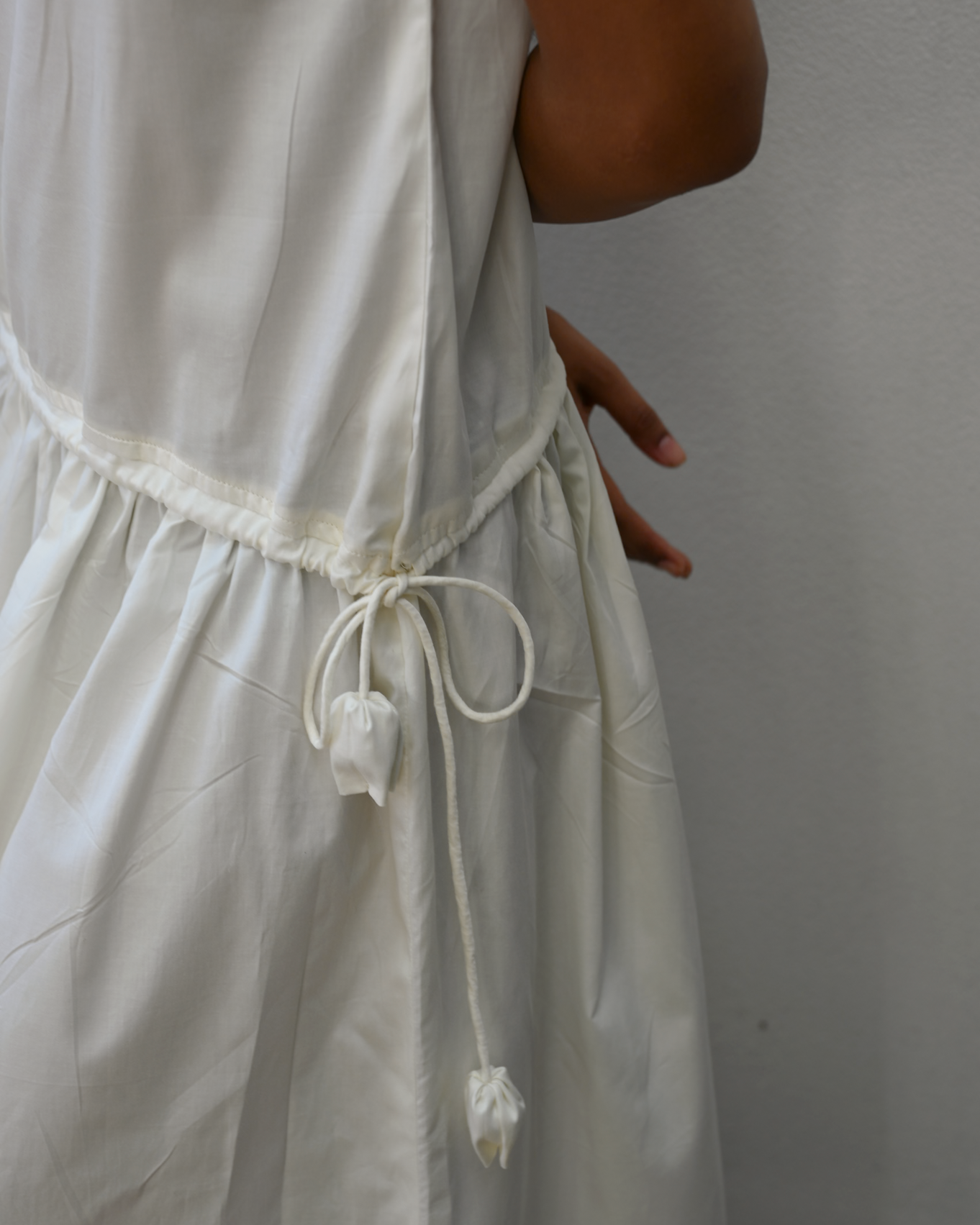 Close-up of a person wearing a white sleeveless dress with a drawstring waist, shown against a plain light gray wall.